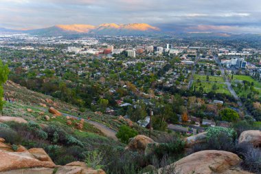 Rubidoux Dağı 'nın yukarıdan görünüşü, sıcak bir günbatımı altında geniş bir nehir kenti manzarası sergiliyor. Uzak dağlar altın ışıkla aydınlatılıyor ve aşağıdaki dolambaçlı patika..