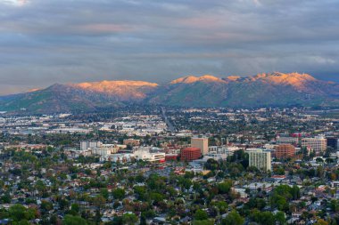 Riverside, Kaliforniya 'nın geniş kentsel yayılımı, altın saatin yumuşak, ılık ışığıyla yıkanmış, bulutlu, atmosferik bir gökyüzü altında, engebeli, güneşli dağ zirveleriyle çevrelenmiştir..