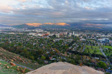 Riverside, Kaliforniya 'nın genişleyen şehir ve yerleşim alanları Rubidoux Dağı' ndan görüldüğü gibi dramatik ve bulutlarla dolu bir gökyüzünün altındaki uzak, güneşli dağlara doğru uzanıyor..