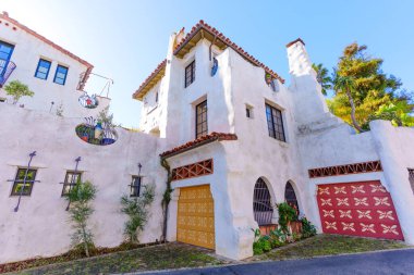 Beyaz sıva, kırmızı kiremit çatılar, kemerli pencereler ve berrak gökyüzü altında parlak desenli garaj kapıları (altın ve kırmızı) bulunan güzel Santa Barbara konutları.