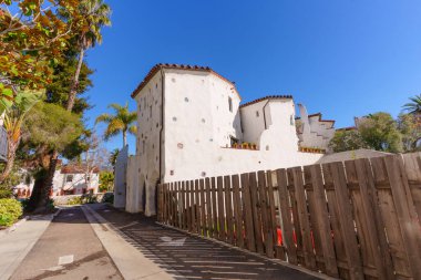 Beyaz sıva, kırmızı kiremit çatı ve dekoratif yuvarlak pencere aksanlı eşsiz Santa Barbara binası, kırsal bir ahşap çitin yanındaki bir ara sokaktan görülüyor..