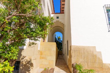Beyaz sıva binaları ve altın taş istinat duvarları tarafından çerçevelenmiş, dar, taştan kemerli geçit, palmiye ağaçlarının ve mavi gökyüzünün parlaklığına yol açıyor..