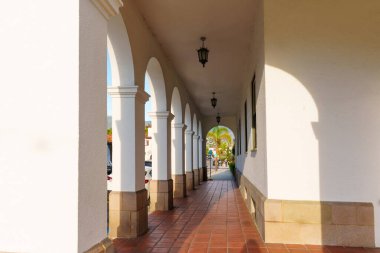 Huzur dolu, güneşli koridorda, sıcak terasotta fayanslarının üzerine gölgeler düşüren beyaz alçı kemerler ve sütunlar serisi var..