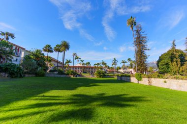 Santa Barbara, California - 29 Aralık 2025: Uzun palmiye ağaçlarının gölgeli manikürlü yeşil çimen canlı mavi gökyüzü altında büyüleyici misyon tarzı binalara doğru yol alıyor.