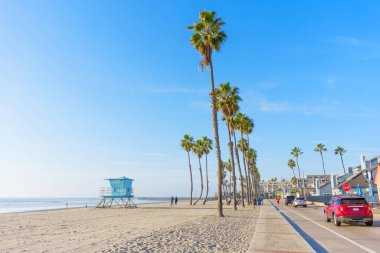 Okyanus Kenarı, Kaliforniya - 17 Aralık 2025 Oceanside 'ın güzel plajının güneşli manzarası, göze çarpan mavi bir cankurtaran kulesi, palmiye ağaçları ve arabalarla dolu hareketli bir kıyı yolu tarafından vurgulandı..