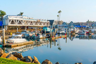 Okyanus Kenarı, Kaliforniya - 17 Aralık 2025: Okyanus kıyısına bakan geniş pencereli modern liman restoranı, çeşitli teknelerin kenetlendiği, kayalık, çimenli bir ön planla çerçevelendiği yer.