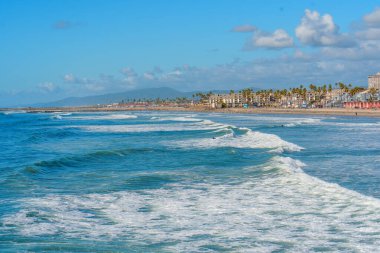 Okyanus Kenarı, Kaliforniya - 28 Aralık 2025: Geniş okyanus manzaralı, California 'nın canlı sahili, uzak sahil binaları ve rıhtımlarıyla kumlara yuvarlanan hafif dalgalar sergiliyor..