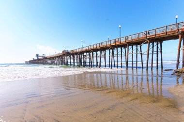 Kaliforniya 'daki Okyanus Kıyısı, Pasifik Okyanusu üzerinde berrak mavi bir gökyüzünün altında uzanır. Sahil sahilinin ıslak kumlarına yansıyan ahşap sütunlar bulunur..