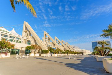 San Diego Kongre Merkezi dış mimarisi ikonik üçgen beton destekleri ve mavi gökyüzünün altında beyaz masalı güneşli bir açık hava plazası sergiliyor..