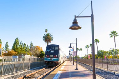 Perris, California - 6 Aralık 2025: Metrolink banliyö treni açık mavi gökyüzünün altında Perris istasyonuna yanaşıyor, palmiye ağaçları ve modern toplu taşıma altyapısı yer alıyor.