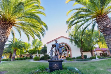 Perris, California - 6 Aralık 2025: Cesar E. Chavez 'in bronz anıtı ve çocuklar Perris, Kaliforniya' daki palmiye ağaçları tarafından çerçevelenmiş şehir kütüphanesi binasının önünde duruyorlar..