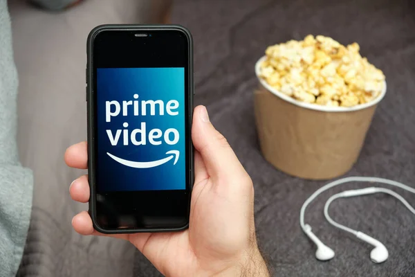 Close up mans hand holding a mobile phone with Prime Video Amazon logo with Apple earphones and popcorn box next him, August 2020, San Francisco, USA — Stockfoto