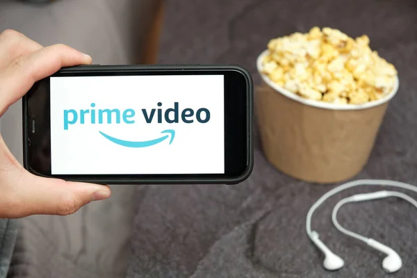 Close up mans hand holding a mobile phone with Prime Video Amazon logo with Apple earphones and popcorn box next him, August 2020, San Francisco, USA — Stockfoto