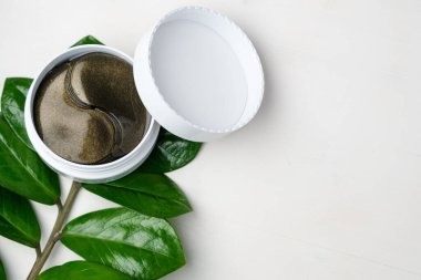 Kozmetik hidrojel göz bantları ve yeşil dallı beyaz arka planda fotokopi alanı olan yapraklar. Cilt bakımı ve kozmetoloji konsepti. Kırışıklık önleyici kolajen maskesi kaldırılıyor.