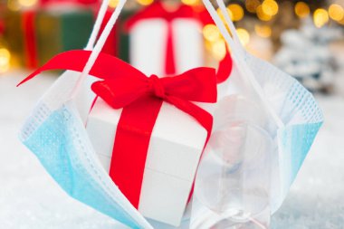 Kurdeleden kırmızı bir fiyonk hediye kutusu, koronavirüs için koruyucu yüz maskesi takılı dezenfektan şişesi Noel ağacı altındaki hediyelerin arka planında. Noel ve Yeni Yıl tatilleri Covid-19 salgını sırasında. 