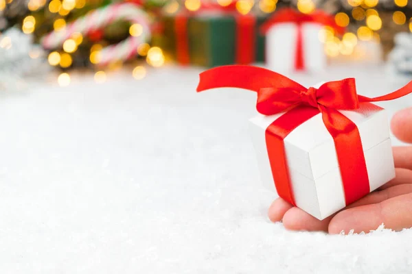 Close up woman hand holding a white gift box with a red ribbon on the background of presents, candy stick, Christmas tree and golden lights with copy space Stock Picture