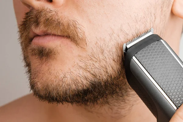 Мужчина бреет бороду и щеки электрической бритвой. Утренняя программа. Обрезка бороды с триммером. — стоковое фото