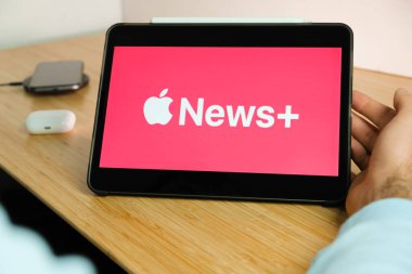 Apple News artı logosu iPad tabletinin ekranında akıllı telefon iPhone 'u kablosuz şarj aleti ve hava podları ile arka planda, Kasım 2020, San Francisco, ABD.