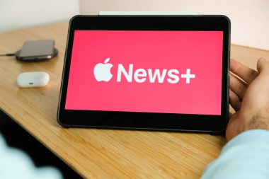 Apple News artı logosu iPad tabletinin ekranında akıllı telefon iPhone 'u kablosuz şarj aleti ve hava podları ile arka planda, Kasım 2020, San Francisco, ABD
