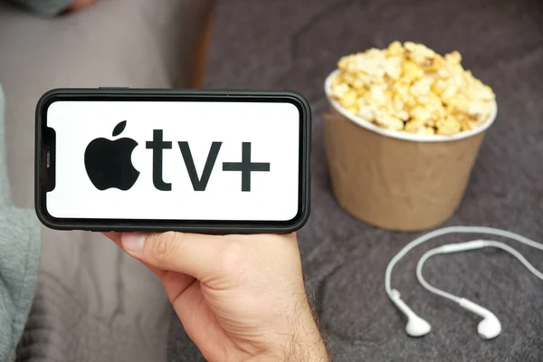 In Großaufnahme hält seine Hand ein Handy mit Apple TV plus Logo mit Apple-Kopfhörern und Popcorn-Box neben sich, August 2020, San Francisco, USA — Stockfoto
