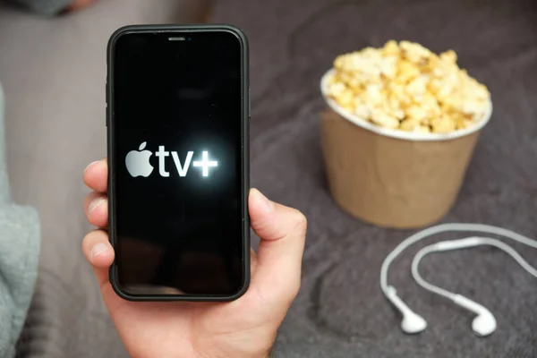 In Großaufnahme hält seine Hand ein Handy mit Apple TV plus Logo mit Apple-Kopfhörern und Popcorn-Box neben sich, August 2020, San Francisco, USA — Stockfoto