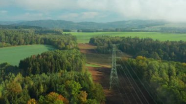 Yüksek voltajlı elektrik hatları ve yüksek voltajlı elektrik iletim kulesi ya da güneş ışığı altında ağaçlarla çevrili bulutların arasındaki pilon boyunca uçmak.
