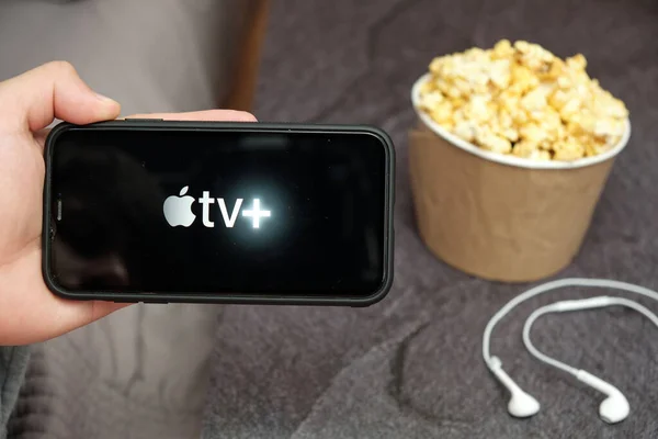 In Großaufnahme hält seine Hand ein Handy mit Apple TV plus Logo mit Apple-Kopfhörern und Popcorn-Box neben sich, August 2020, San Francisco, USA — Stockfoto