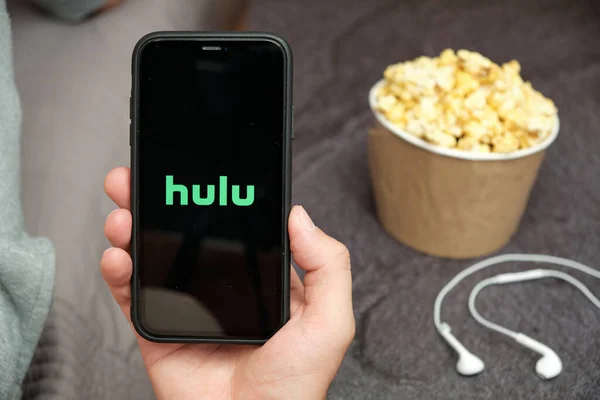 Close Mans hand holding a mobile phone with Hulu logo with Apple earphones and popcorn box next on him, sierpień 2020, San Francisco, USA — Zdjęcie stockowe