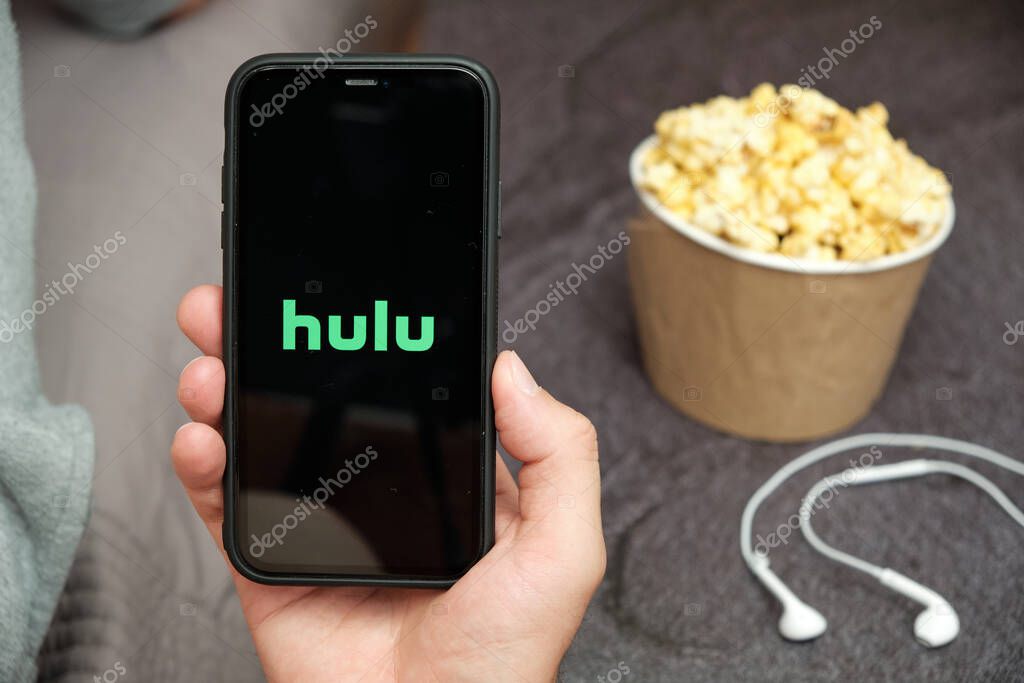 Close up mans hand holding a mobile phone with Hulu logo with Apple earphones and popcorn box next to him, August 2020, San Francisco, USA.