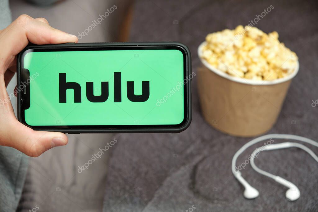Close up mans hand holding a mobile phone with Hulu logo with Apple earphones and popcorn box next to him, August 2020, San Francisco, USA.