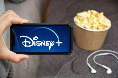 Yanında Disney logosu, Apple kulaklıkları ve patlamış mısır kutusu olan bir cep telefonu olan bir adamın elini kapat, Ağustos 2020, San Francisco, ABD