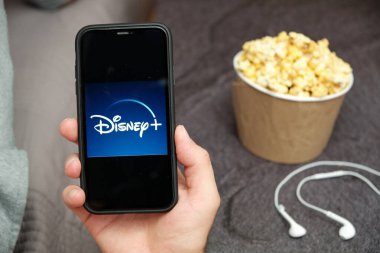 Yanında Disney logosu, Apple kulaklıkları ve patlamış mısır kutusu olan bir cep telefonu olan bir adamın elini kapat, Ağustos 2020, San Francisco, ABD