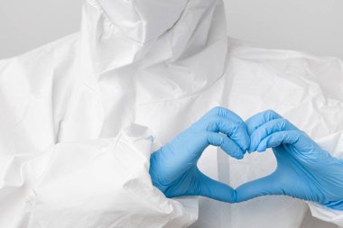 Koronavirüs, Sars-Cov-2 ve Covid-19 'a karşı koruyucu koruyucu PPE kıyafeti ve koruyucu yüz maskesi takmış bir doktor mavi lastik eldivenlerle kalp şeklinde el ele tutuşuyor..