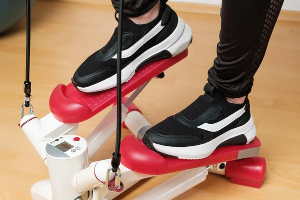 Close up womans feet in sneakers training on the twist stepper with digital display and gum expanders at home during lockdown.