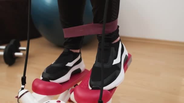 Sluiten vrouw voeten doen cardio op de stepper en met behulp van elastische banden met bal, en halters op de achtergrond. Workout thuis. — Stockvideo