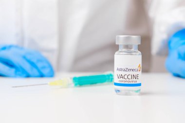 Koronavirüse karşı Astra Zeneca aşısı ve enjeksiyon için şırınga masada ve sağlık çalışanı elinde kauçuk eldivenler ve koruyucu giysi ile arka planda, Mart 2021, San Francisco, ABD