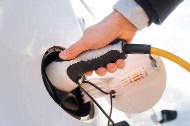 Takım elbiseli adam güneş ışığında caddedeki şarj istasyonunda elektrikli bir arabaya güç kaynağı takıyor. 