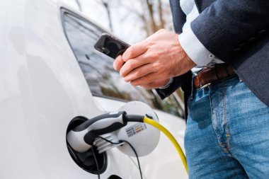 Elektrikli arabanın yanında duran takım elbiseli bir adamı kapat ve akıllı telefonunu para ödemek için kullan. 