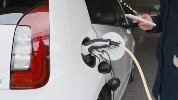 Γυναίκα αποσυνδέοντας το καλώδιο τροφοδοσίας ρεύματος από ένα ηλεκτρικό αυτοκίνητο και χρησιμοποιώντας το smartphone της για το ξεκλείδωμα του οχήματος — Αρχείο Βίντεο