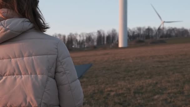 Kvinnlig ingenjör i orange vesta går till vindkraftverk med en surfplatta för att kontrollera deras drift. — Stockvideo