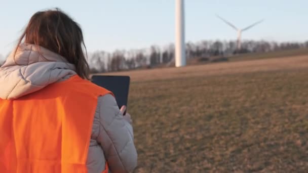 Kvinnlig ingenjör i orange vesta går till vindkraftverk med en surfplatta för att kontrollera deras drift. — Stockvideo