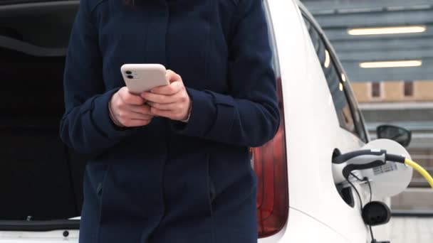 Mulher de pé perto do tronco usando seu telefone celular e esperando pelo carregamento do carro elétrico. — Vídeo de Stock