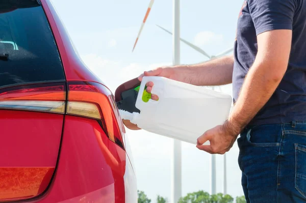 Člověk plní motorovou naftu z kanystru do nádrže červeného auta na pozadí větrných turbín. Snížení znečištění ovzduší a ochrana životního prostředí. Koncept zelené energie. — Stock fotografie