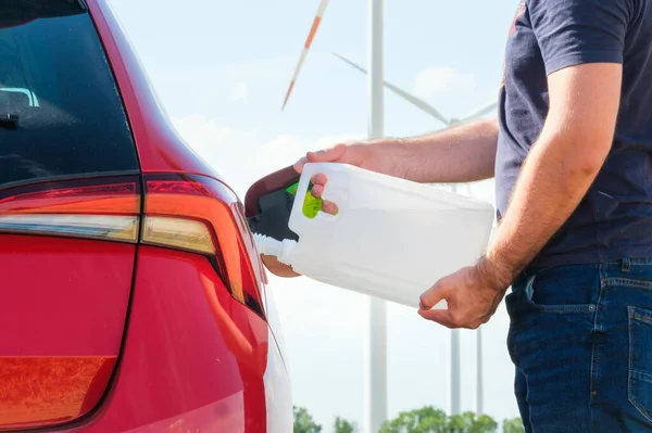 Homem enchendo um fluido do motor diesel da lata para o tanque de carro vermelho no fundo de turbinas eólicas. Redução da poluição atmosférica e protecção do ambiente. Conceito de energia verde. — Fotografia de Stock