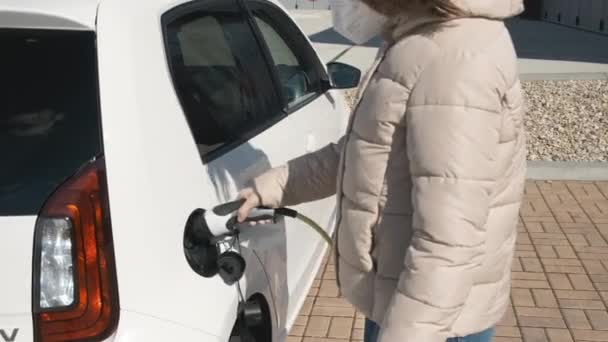 Mujer con mascarilla protectora o respirador enchufando un coche eléctrico para cargar en la calle en el punto de carga. — Vídeo de stock