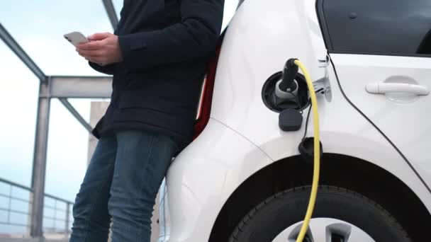 Мужчина, стоящий возле багажника, используя ее мобильный телефон и ожидающий зарядки электромобиля. — стоковое видео