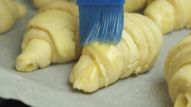 Zbliżyć rozprzestrzenianie żółtka jaj na włoskich lub francuskich rogalików z silikonową szczotką do układania na papierze piekarniczym. Ciasto domowe. — Wideo stockowe