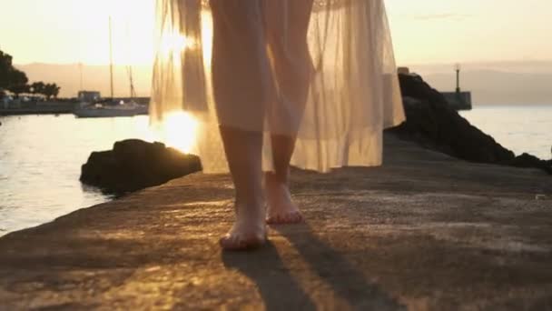 Egy nő sétál a mólón a tenger mentén fehér átlátszó ruhában napkeltekor vagy napnyugtakor.. — Stock videók