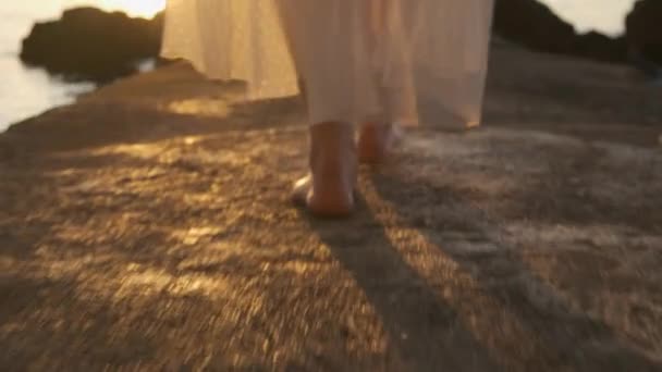 Une femme marche le long de la jetée le long de la mer dans une robe blanche transparente au lever ou au coucher du soleil. — Video