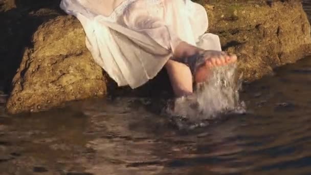 Egy fehér ruhás nő, aki napkeltekor lábakon lóg. Lazító és megható tenger lábával víz a szikláról naplementekor. — Stock videók